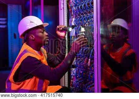 Concentrated Black workman in hardhat and orange vest crouching by server and using flashlight while fixing server problem