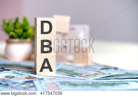 Wooden Cubes With The Letters Dba Arranged In A Vertical Pyramid On Banknotes, Green Plant In A Flow