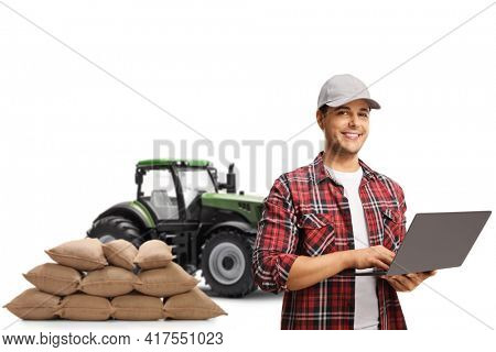 Farmer with a cap and a checkered shirt working on a laptop computer and standing in front of a tractor isolated on white background