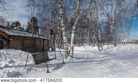 On The Ground Covered With Snow, Paths Are Trodden, Nearby Are Snowdrifts. There Is A Wooden Swing -
