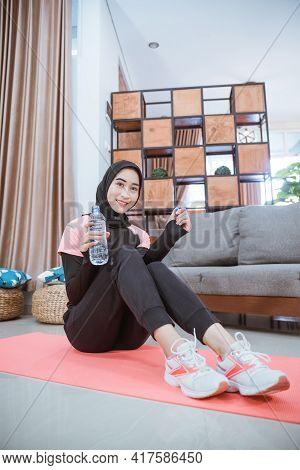Shot From Below Of Muslim Woman Wearing A Hijab Gym Outfit Sits On The Floor Holding A Drinking Wate