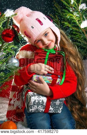 A Baby Girl In A Red Jacket And A Cap With A Pumpon, Hugs Gifts. Happy To Receive Gifts. Joy Of New 