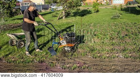 Male In The Spring Plows The Earth In Working Clothes With A Motoblock, Walk-behind Tractor