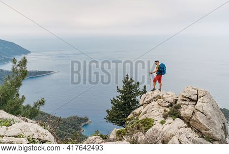 Young Hiker Backpacker Man Using Trekking Poles Enjoying The Mediterranean Sea On Clifftop During Ly