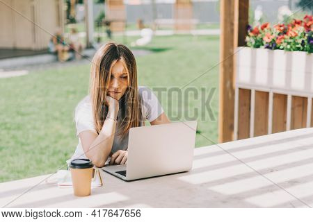 Beautiful Sad Thinking Woman Working At Laptop Outside And Drinking Coffee. Using Computer And Onlin