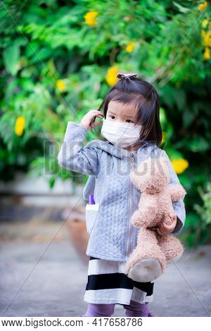 Adorable Girl Wearing Medical Face Mask Holds Brown Teddy Bear Walking In Front Yard. Prevent The Sp