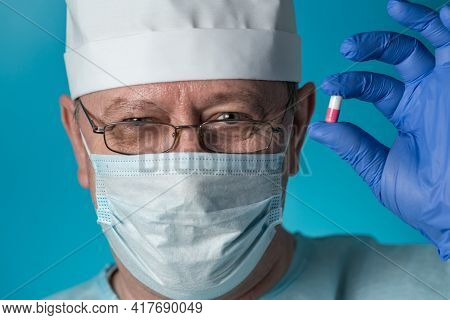 An Elderly Doctor In Medical Clothing: A Cap, Gloves,dressing Gown And Mask, Glasses. He Holds A Med