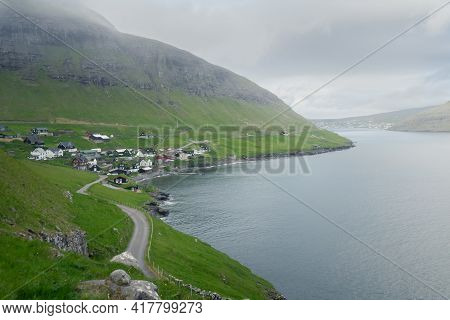 A Road Heading To Faroese Village Of Bour On A Cold And Cloudy Day. Green Grass On Steep Cliffs And 