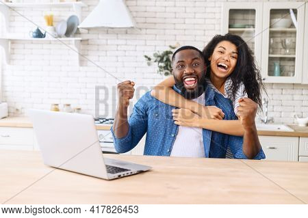 Overjoyed Multiracial Couple In Love In The Kitchen At Home Celebrating Victory, Sits In Embrases In