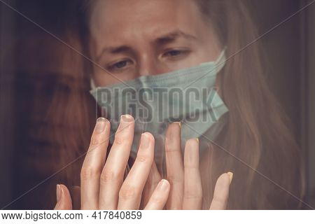 Isolation Of The Coronavirus. Hapless Dark-haired Girl In A Mask Touches A Woman's Hand Through A Gl
