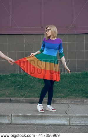 The Girl Stands In Glasses And A Colored Dress, Looks Aside At The One Who Holds And Pulls The Edge 