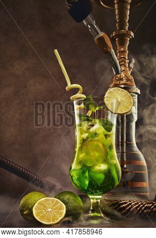 Hookah (shisha) and glass of Mojito cocktail with lime and ice on dark brown wooden table on dark background with smoke. Weekend or holiday party