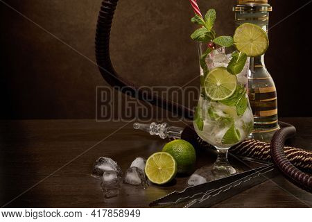 Hookah (shisha) and glass of Mojito cocktail with lime and ice on dark brown wooden table on dark background. Weekend or holiday party