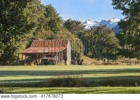 Old Red Farm Shed With Disused Tractors Under Lean-too With Snow-capped Mountains In Background. In 