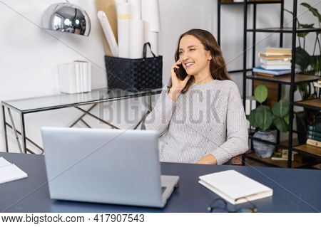 Positive Young Female Entrepreneur In Casual Wear Talking On The Smartphone Sitting At The Desk, Che