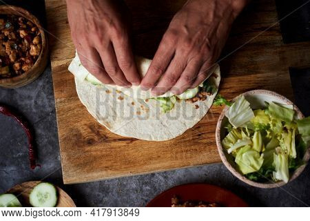 a young caucasian man is rolling a durum or a burrito, filled with chicken meat cooked with onion and red and green pepper, and fresh lettuce and raw cucumber