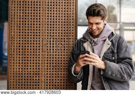 Smiling young white man in casual cothes using mobile phone outdoors in the city, leaning on a wall