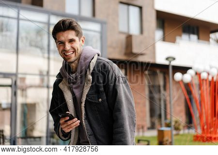 Smiling young white man in casual cothes using mobile phone outdoors in the city, walking