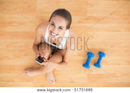 Smiling sporty brunette holding a mobile phone and sitting next to dumbbells on the floor