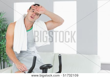 Exhausted handsome man exercising on exercise bike in bright living room
