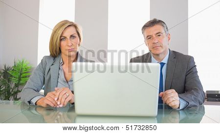 Two severe mature business people looking at camera behind an opened laptop at office