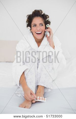 Laughing natural brunette applying nail polish and making a call in bedroom
