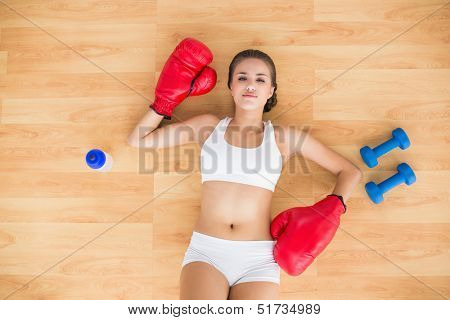 Tired brunette wearing red boxing gloves and lying next to a water bottle and dumbbells on the floor