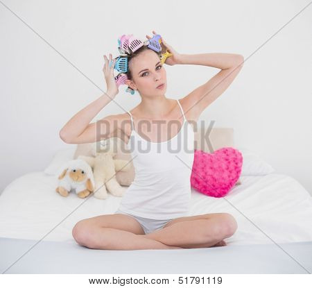 Pouting natural brown haired woman fixing her hair curlers in bright bedroom