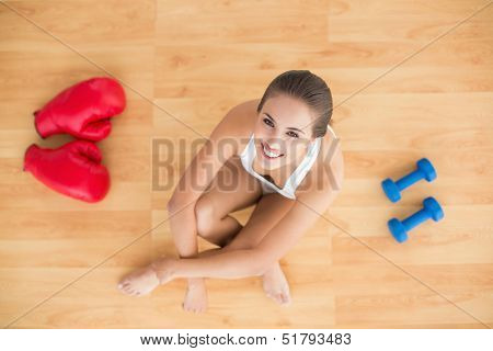 Sporty brunette woman sitting next to red boxing gloves and dumbbells on the floor