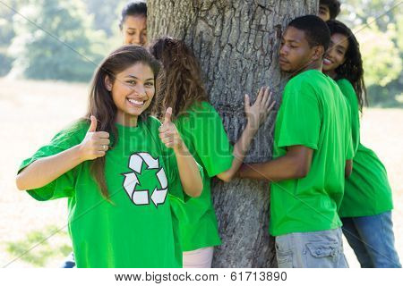 Portrait of confident female environmentalist showing thumbs up with friends hugging tree in background