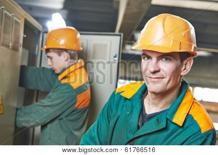 Happy young adult electrician builder engineer in front of his co-worker screwing equipment in fuseboard