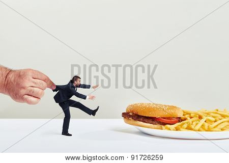 small man running to junk food but the big hand holding him over light grey background