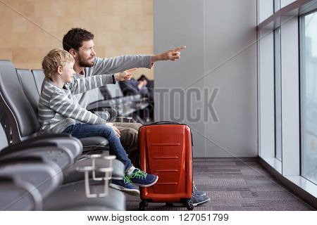 positive smiling boy and his father waiting at the airport for plane departure and pointing with fingers at something vacation and travel concept