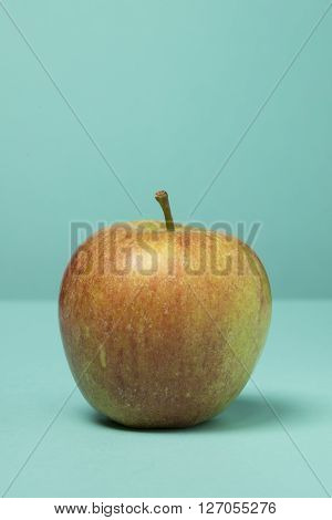 A single red cox apple on a blue green background lit from the left with a soft shadow to side