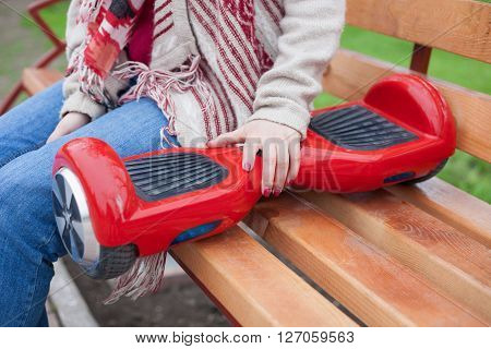 Girl in boho style outfit sitting on a bench with a modern red electric mini segway or hover board scooter. Trending new transportation technology that is so much fun and easy to ride and produces no air pollution to the atmosphere. Focus on the device