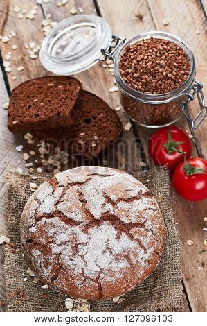 Closeup photo of rustic floured bread on sackcloth, two pieces of brown tommy, tomatoes, oat flakes and jar with buckwheat on planked wooden table