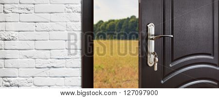 Entrance door half opened to nature background. Door handle, door lock. Exit outdoors. Opening door. Entrance to the house. Door at white brick wall, summer field and forest, freedom concept.