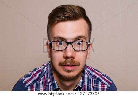 Face of the surprised man. A young man in glasses and a shirt with a surprised look into the camera. Guy with wide open eyes with glasses a beard and mustache.