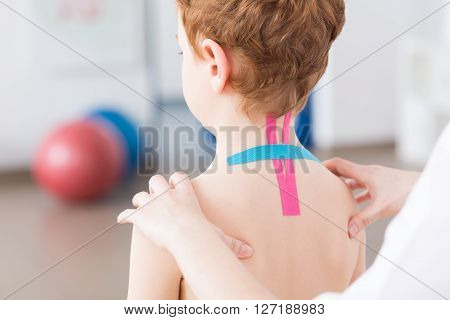 Little crooked boy with spine problems having kinesiotaping on his cervical spine. Physiotherapist exercising with little patient