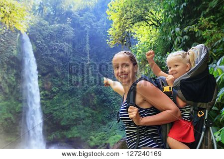 Mother hold baby girl on back in carrying backpack walking in waterfall canyon. Travel adventure and hiking activity with child active and healthy lifestyle on family summer vacation and weekend tour