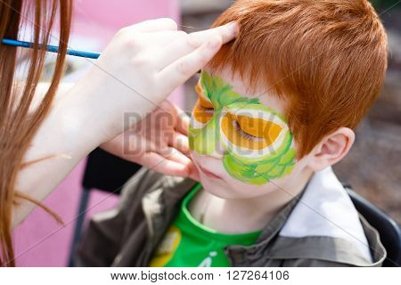 Child animator, artist's hand draws face painting to redhead child. Redhead boy with funny face painting. Painter makes reptile eyes at him. Children holiday, event, bithday party, entertainment.