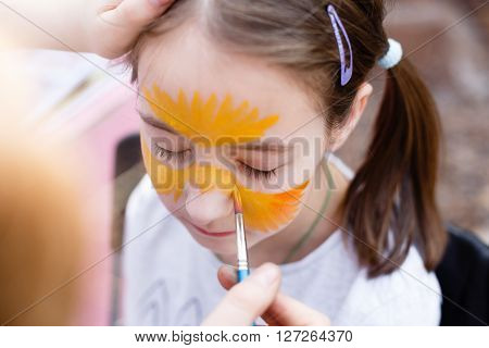 Child animator, artist's hand draws face painting to little girl. Child with funny face painting. Painter makes tiger eyes at girl's face. Children holiday, event, bithday party, entertainment.