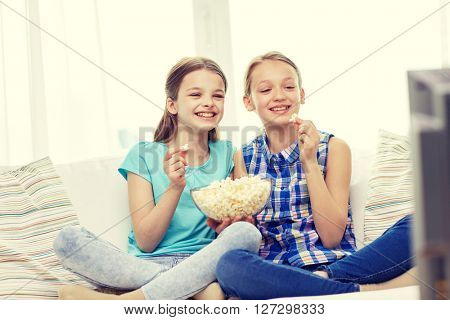 people, children, television, friends and friendship concept - two happy little girls watching comedy movie on tv and eating popcorn at home