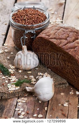 Close up photo of rustic bread on sackcloth, garlic, dill, oat flakes and jar with buckwheat on planked wooden table