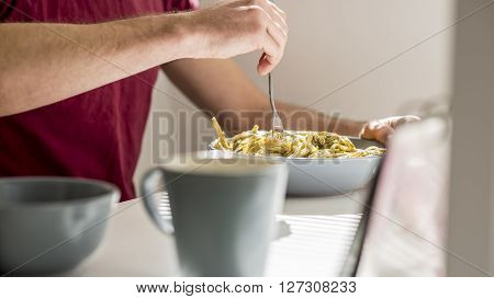 Closeup of a man swirling spaghetti with pesto sauce with a fork sitting at white desk with coffee mug in front of the pasta plate.