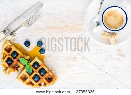 Espresso coffee cup and soft belgian waffles with fresh blueberries and marple syrup on white painted wooden board over light blue background. Top view, copy space, horizontal