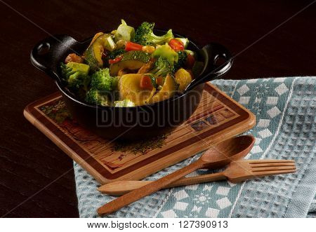 Delicious Homemade Colorful Vegetables Ragout with Zucchini Carrots Broccoli Leek and Red Bell Pepper in Black Iron Stewpot with Wooden Spoon and Fork closeup on Blue Napkin