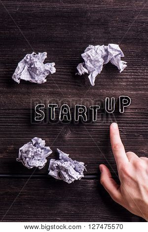 Office desk with start up sign made of cookie cutters and hand of unrecognizable man pointing up. Crumpled paper balls. Flat lay. Workplace. Studio shot on wooden background.