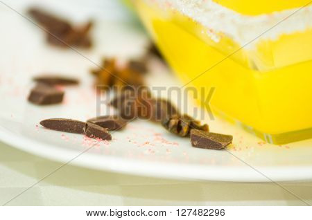 Closeup small pieces of dark chocolate lying on white plate next to transparent square bowl sugarcoated with yellow dessert.