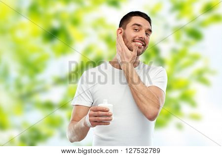 beauty, skin care, body care and people concept - smiling young man applying cream or lotion to face over green natural background